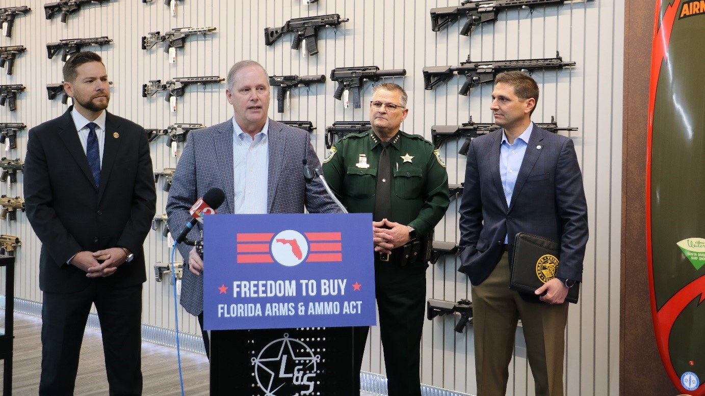 Commissioner Simpson at a podium surrounded by other officials, with guns mounted on the wall behind them